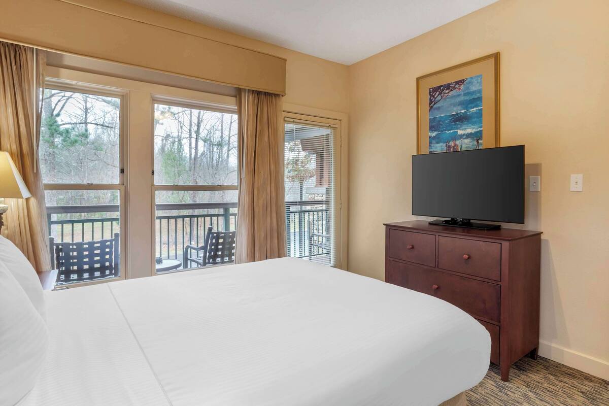A hotel room with a large bed, a television on a wooden dresser, and a balcony with outdoor furniture.