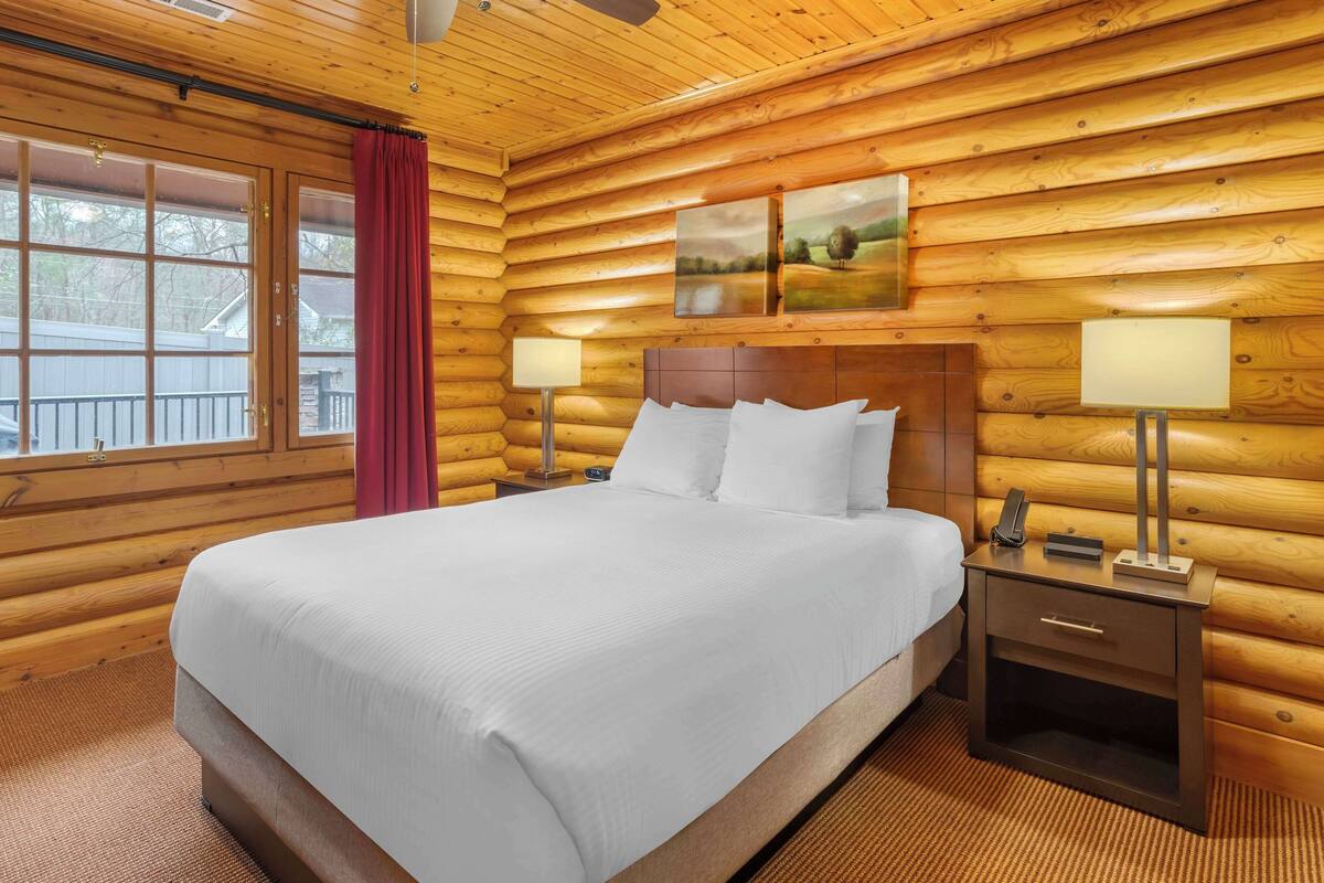 A log cabin bedroom with a large bed, two bedside tables with lamps, and a window with a red curtain.