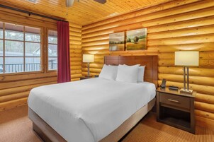 Un dormitorio tipo cabaña de madera con una cama grande, dos mesitas de noche con lámparas y una ventana con cortina roja.