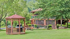 A gazebo with a grill and seating area, a multi-story building with large windows, and a lush green landscape with trees and rocks.