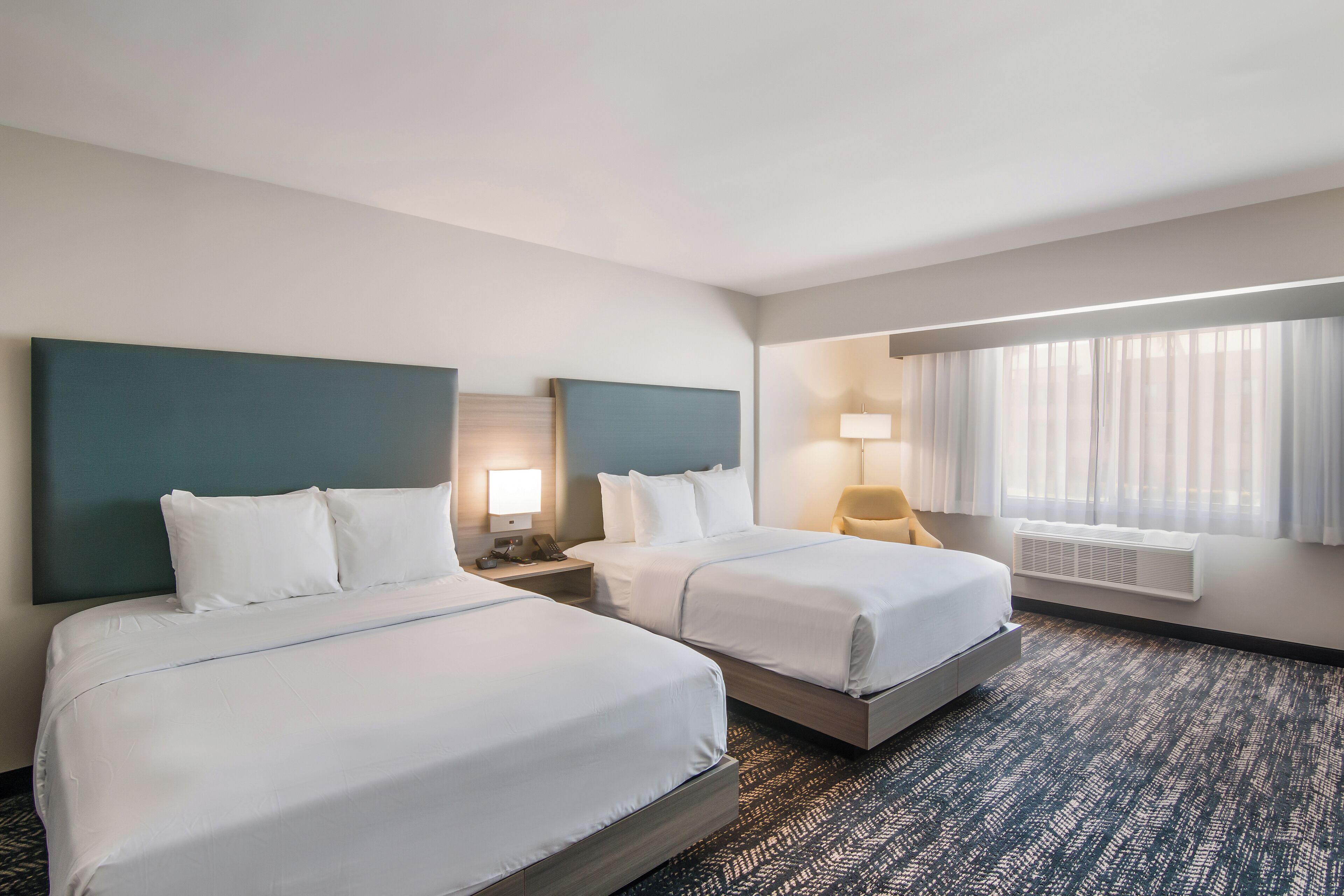 A hotel room with two beds, a large window with curtains, a desk with a lamp, and a wall-mounted air conditioner.