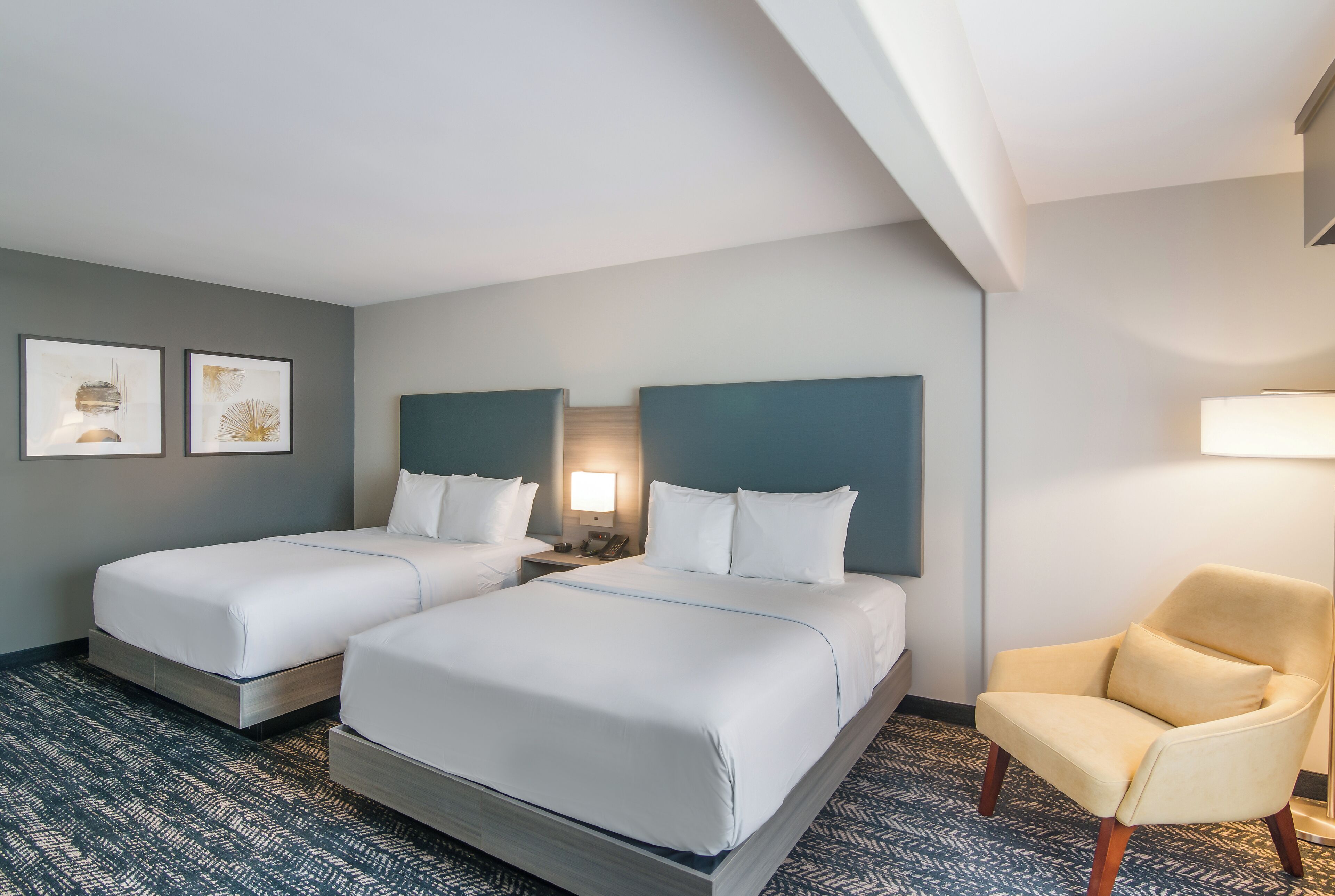 A hotel room with two beds, a chair, and artwork on the wall.
