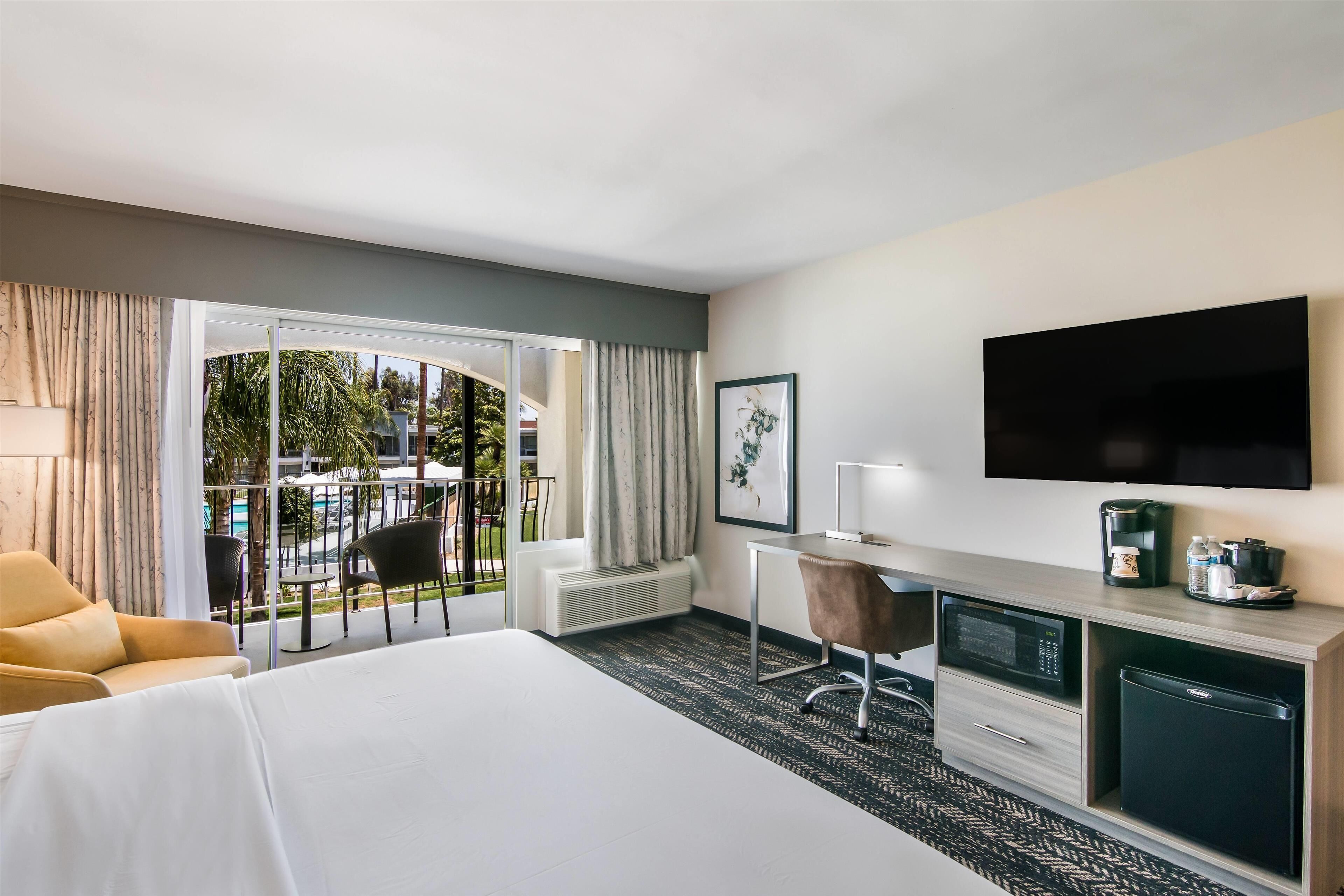 Habitación de hotel con cama, escritorio con silla, televisión, refrigerador pequeño y vista al exterior.