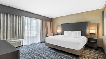 A hotel room with a large bed, a desk, a chair, a nightstand, a lamp, and a window with curtains.