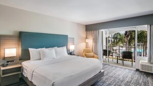 A hotel room with a large bed, a nightstand, a lamp, a chair, and a view of a pool and palm trees through the glass door.