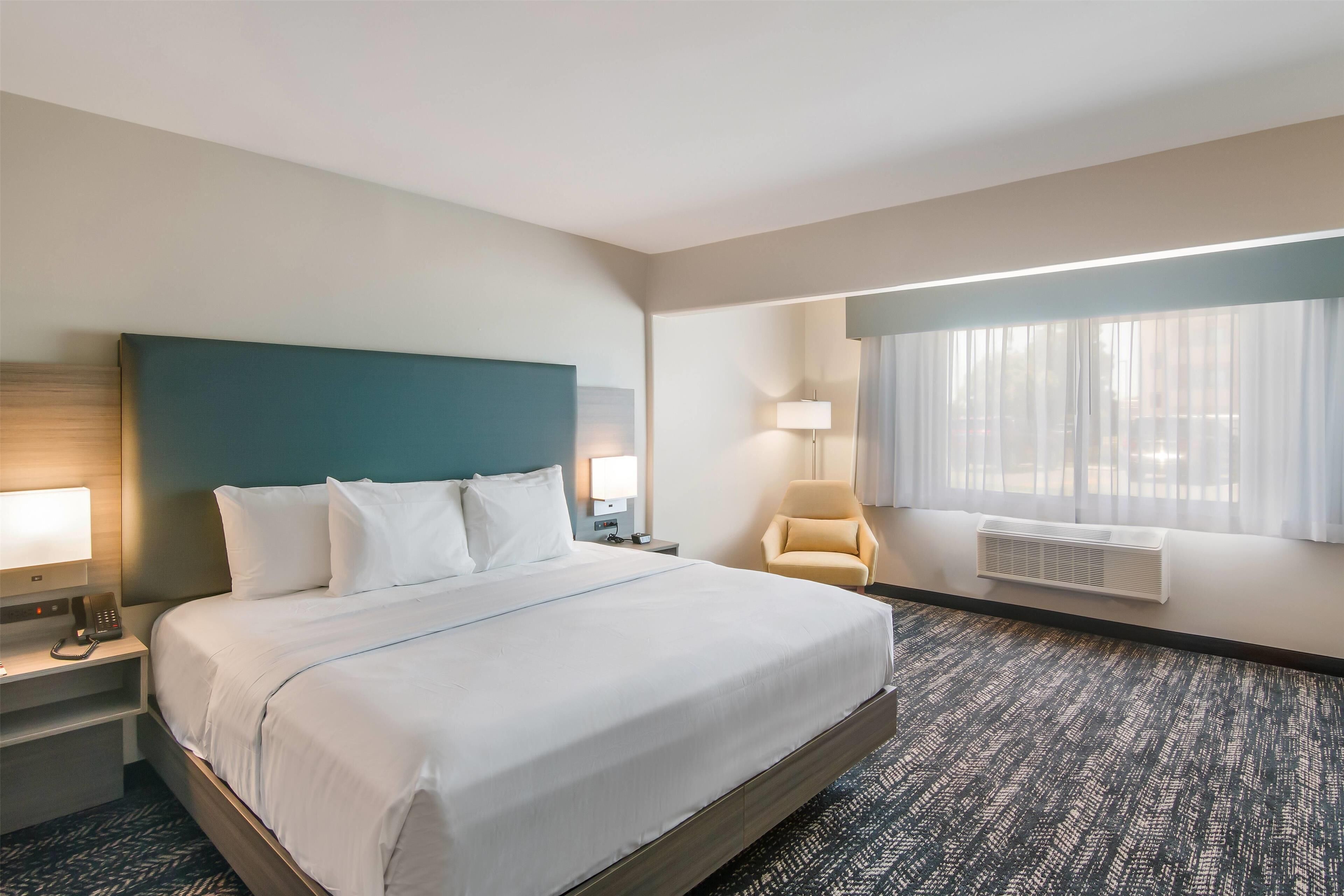 Una habitación de hotel con una cama grande, mesitas de noche con lámparas, una silla, una ventana con cortinas y un aire acondicionado instalado en la pared.