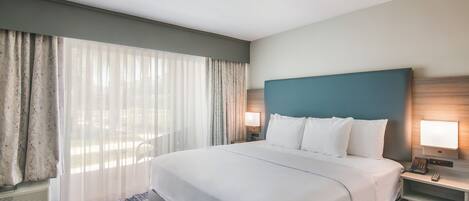 A hotel room with a large bed, two bedside tables with lamps, a window with sheer curtains, and a wall-mounted heater.