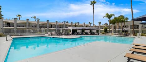 A swimming pool with lounge chairs and umbrellas, surrounded by palm trees and a building with balconies.
