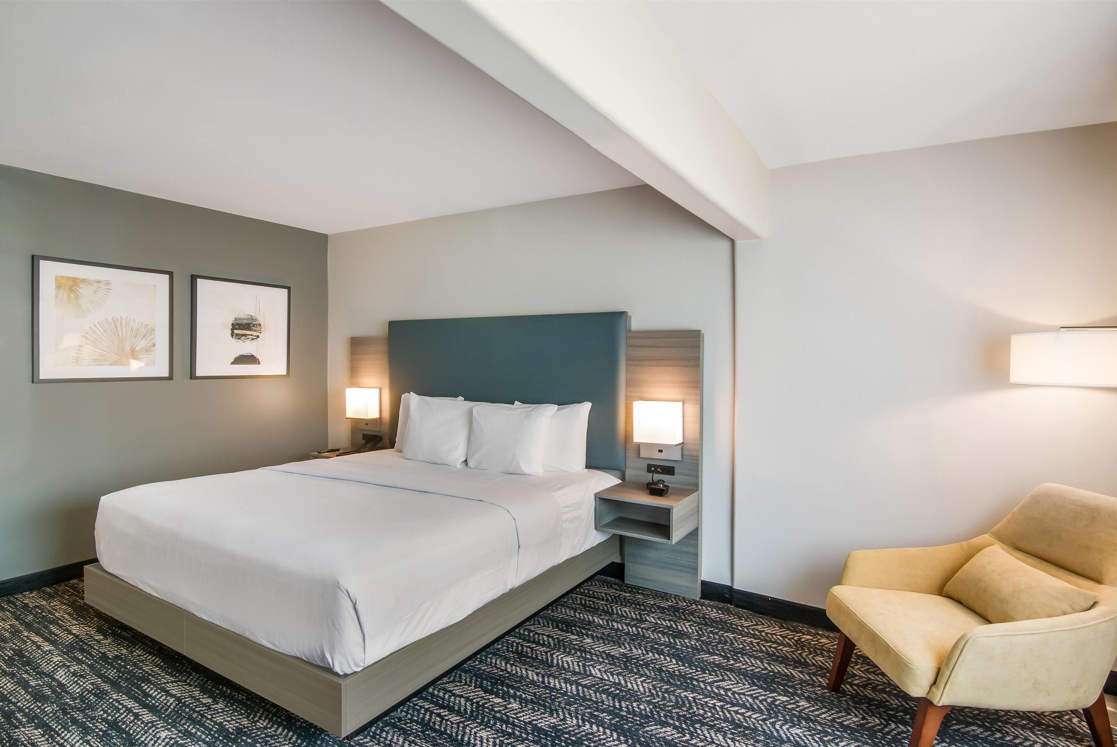 A hotel room with a large bed, two bedside lamps, a headboard, a chair, and two framed pictures on the wall.