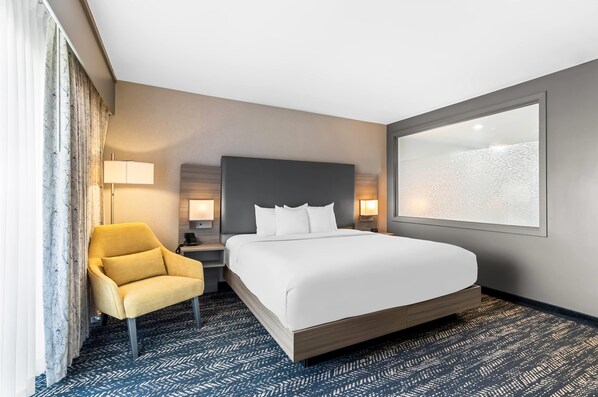 Habitación de hotel con una cama grande, un sillón amarillo, una mesita de noche con lámpara y un ventanal con vistas.