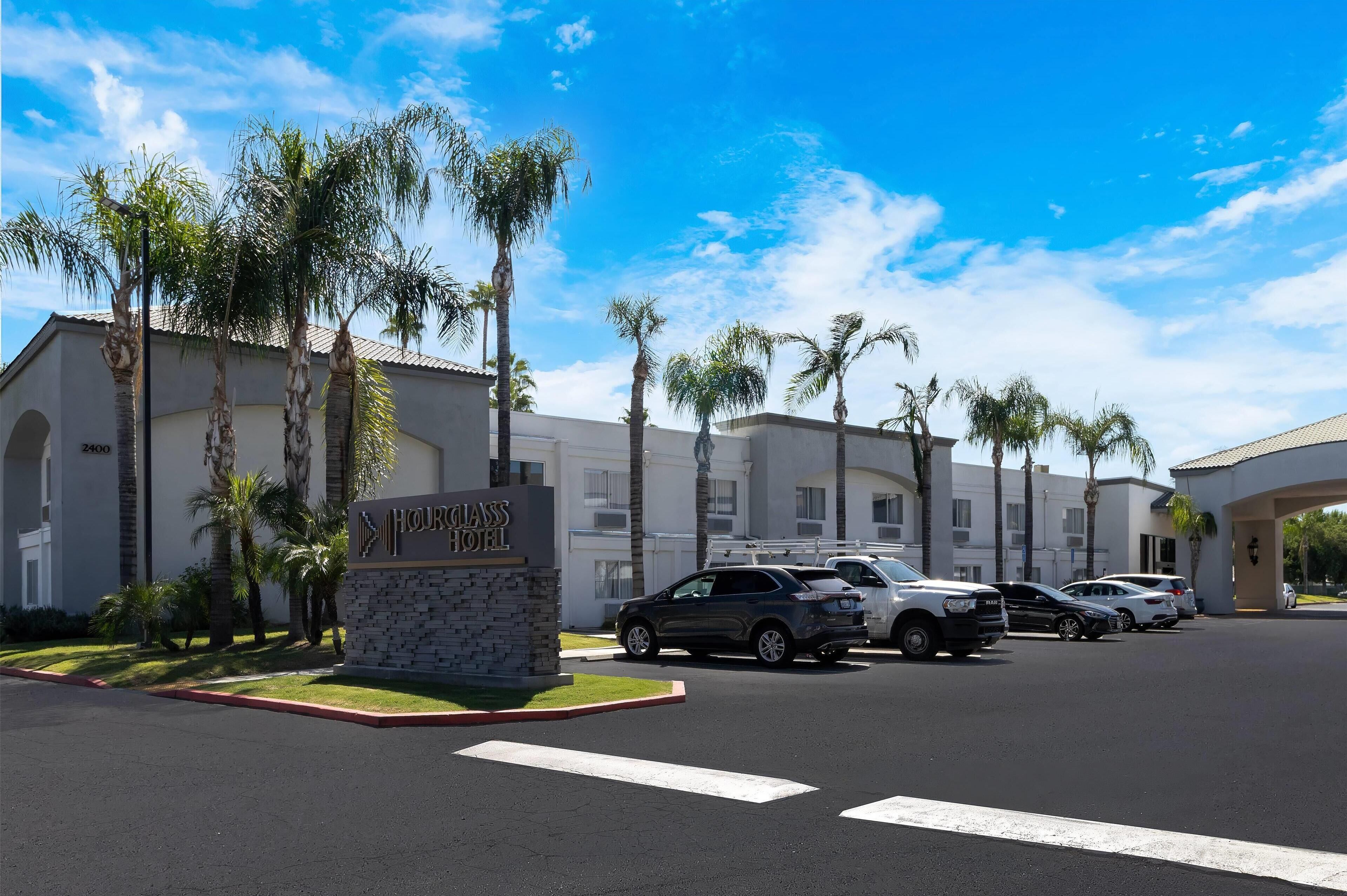 A hotel with a sign reading "Hotel Grand." There are several cars parked in front, palm trees, and a clear blue sky.