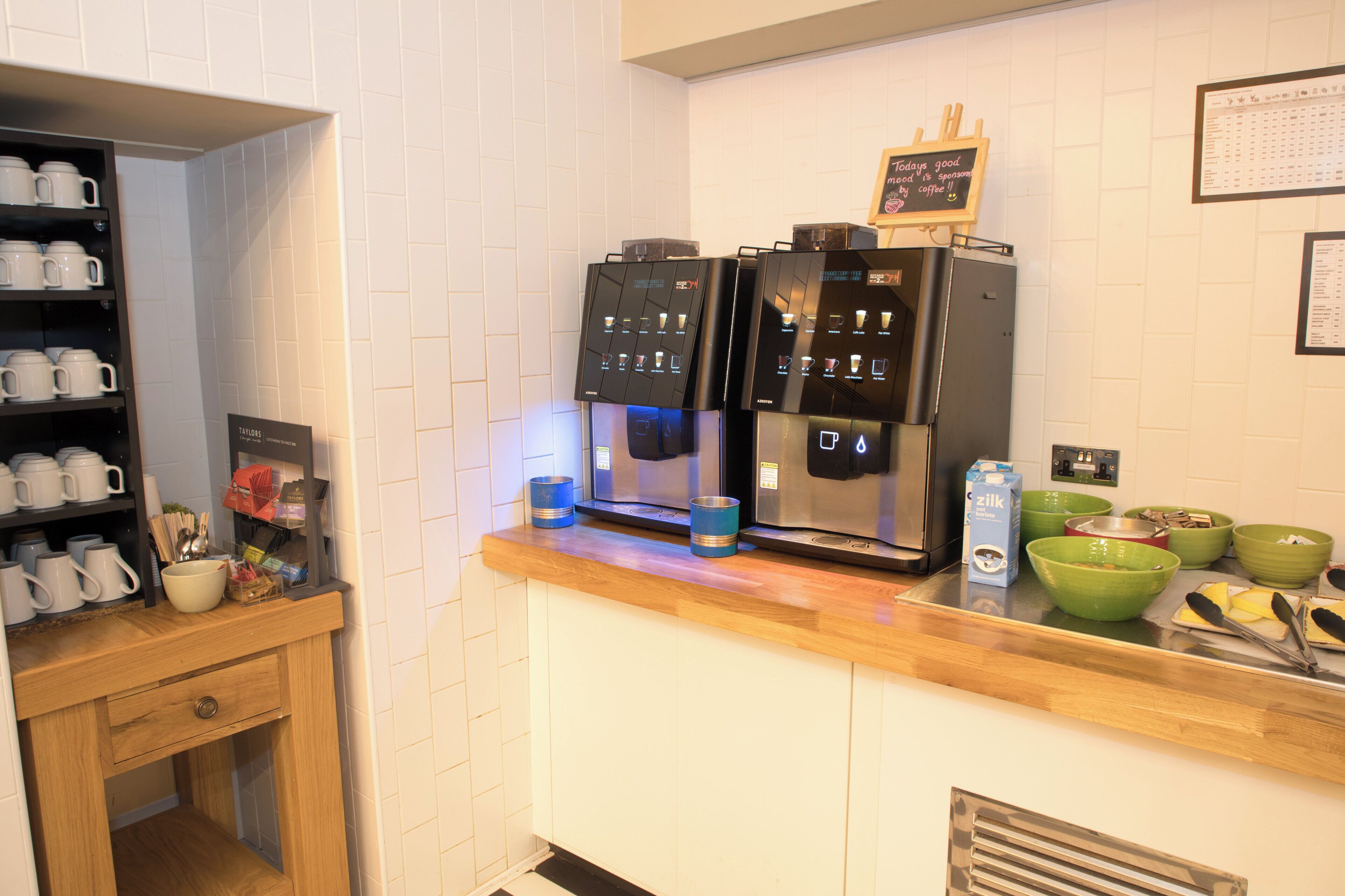 Una cocina con dos máquinas de café, una estantería con tazas y un cartel en la pared.