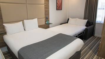A hotel room with two beds, a wooden headboard, a small shelf with amenities, and a window with curtains.