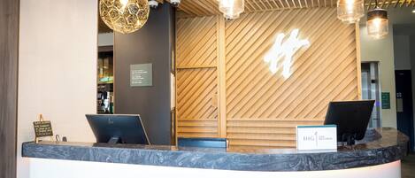 A hotel reception desk with a modern design, including a wooden wall with a geometric pattern, a reception area with two computer monitors, and a sign indicating hotel management.