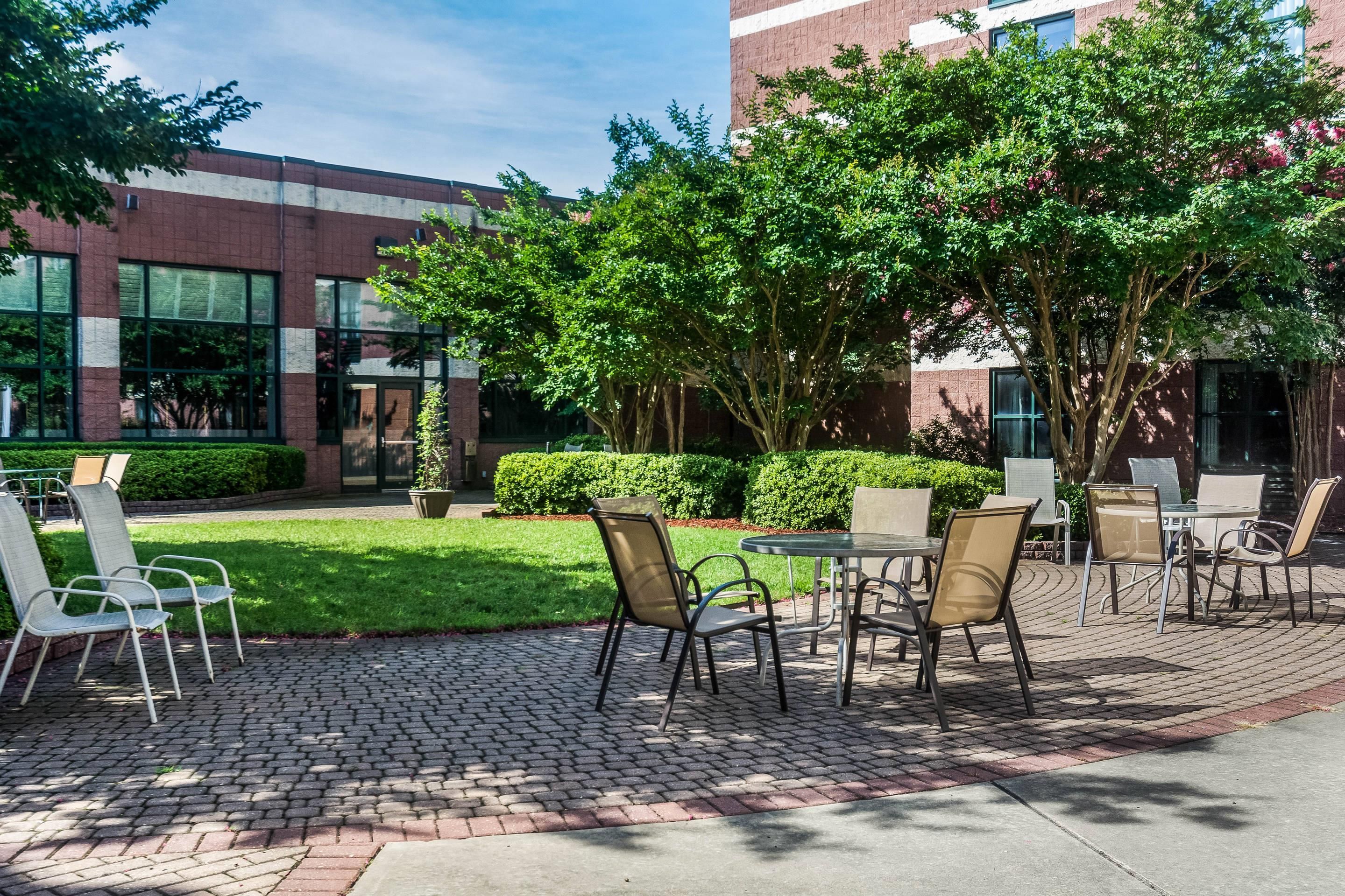 An outdoor seating area with tables and chairs, surrounded by greenery and a brick building.