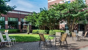 An outdoor patio with brick flooring, metal chairs, and tables, surrounded by greenery and a building with large windows. - Sleep Inn Lake Wright - Norfolk Airport (Norfolk)