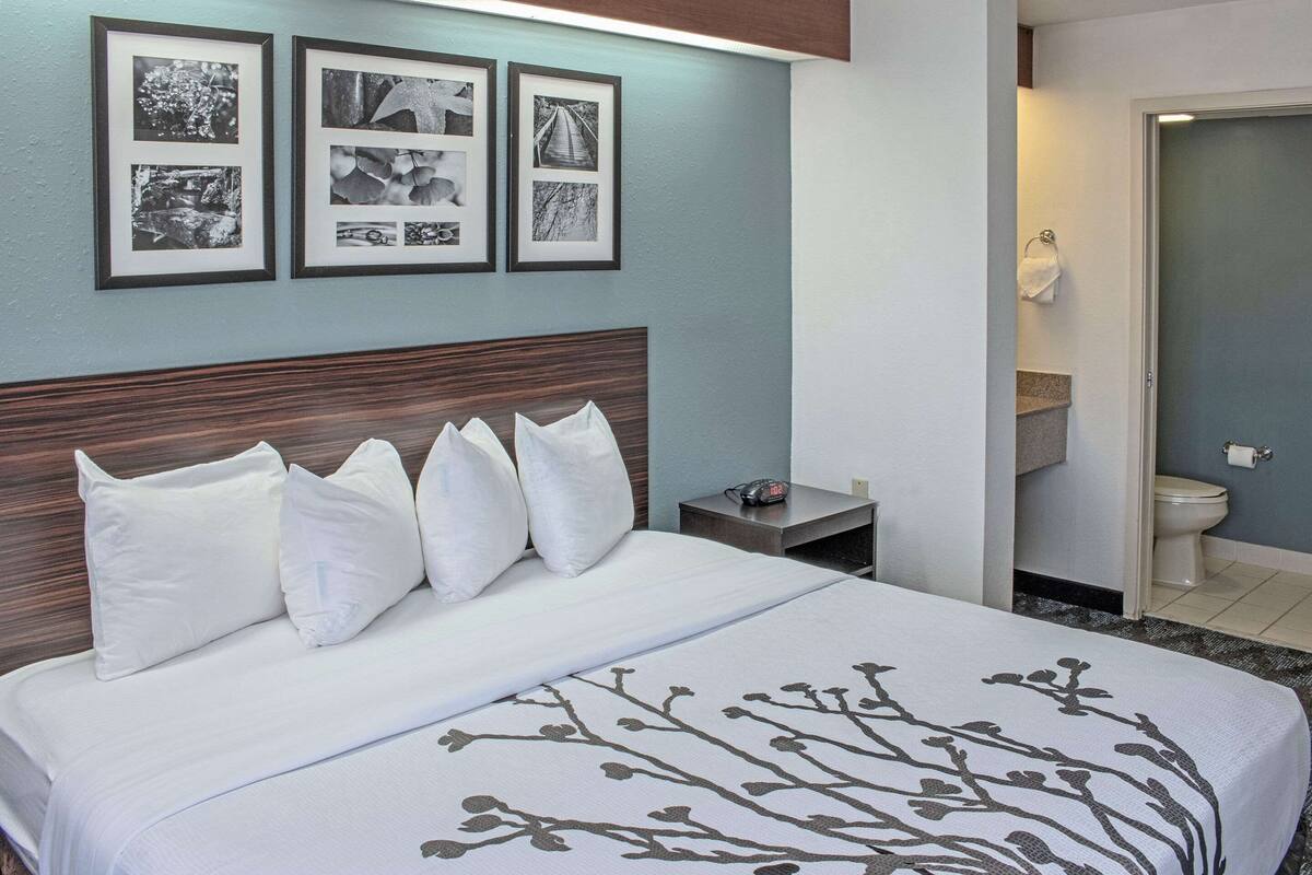 Hotellihuone, jossa on suuri sänky, puinen pääty ja yöpöytä, jossa on kello. Seinällä on kehystettyjä kuvia, ja avoimesta ovesta näkyy kylpyhuone.