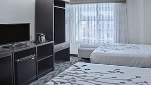A hotel room with a TV, a minibar, a bed with a floral bedspread, and a large window with sheer curtains. - Sleep Inn Lake Wright - Norfolk Airport (Norfolk)