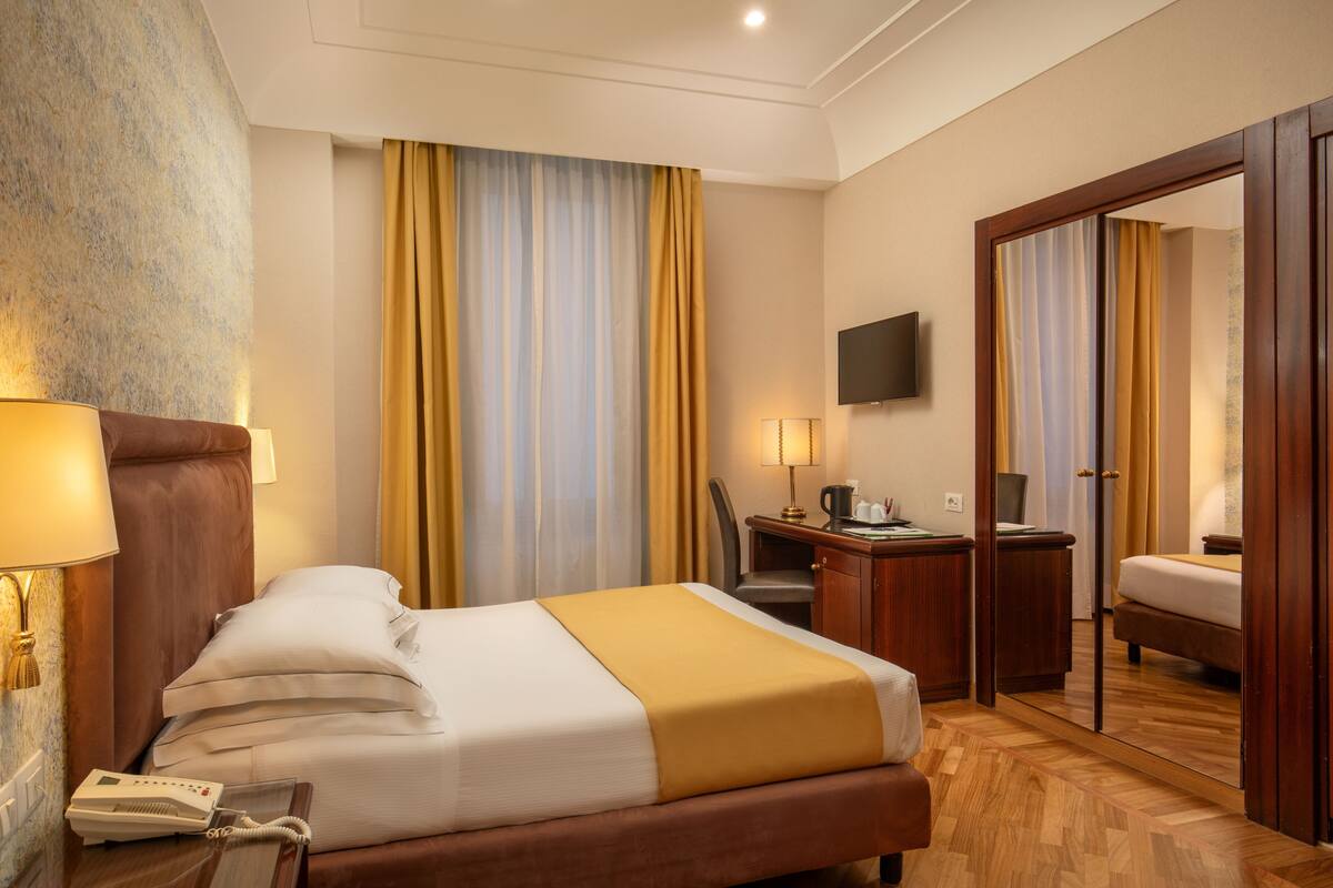 A hotel room with a bed, bedside table, lamp, desk, chair, and a television on the wall.
