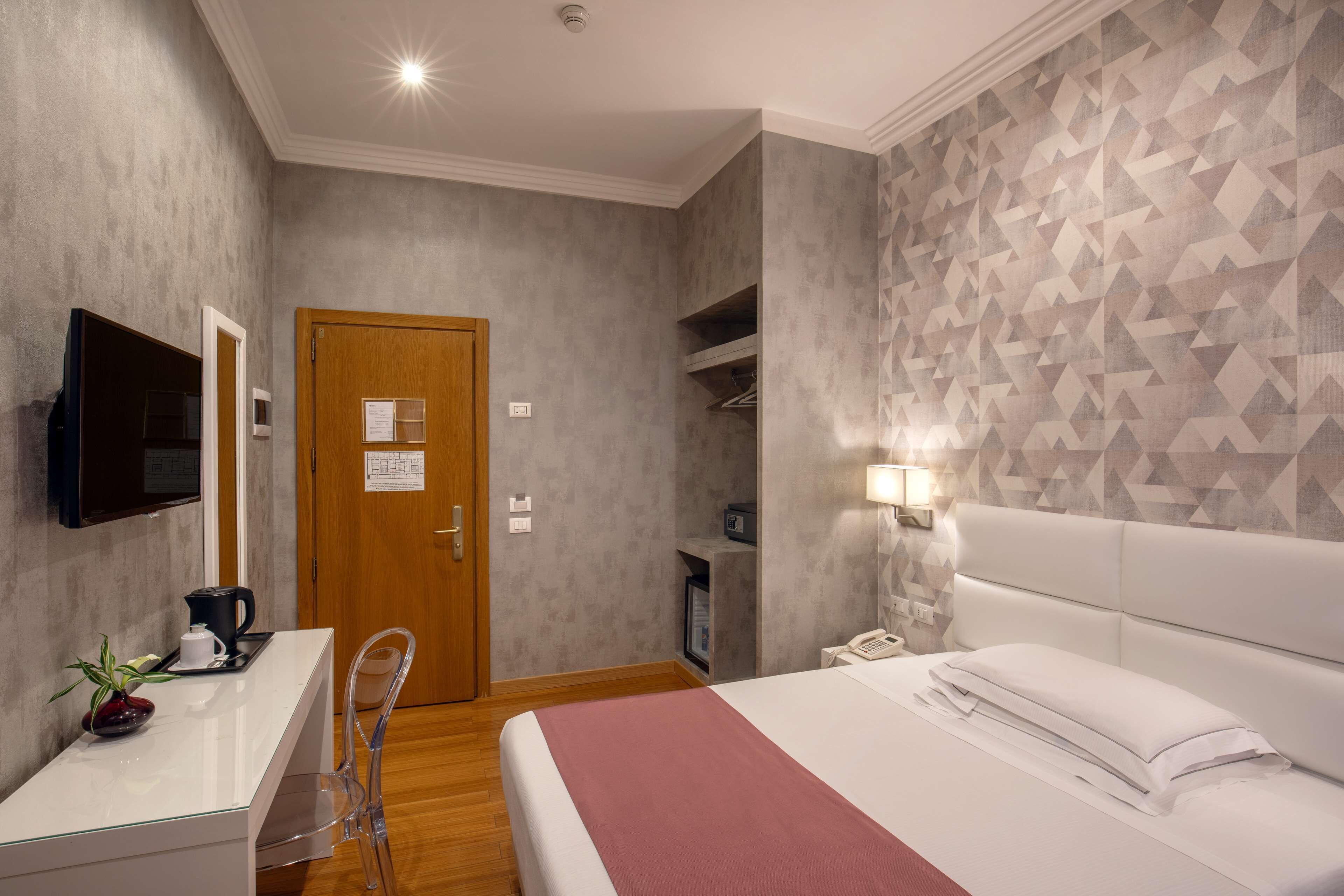 Habitación de hotel con cama, mesita de noche, televisor y una pared con diseño geométrico.