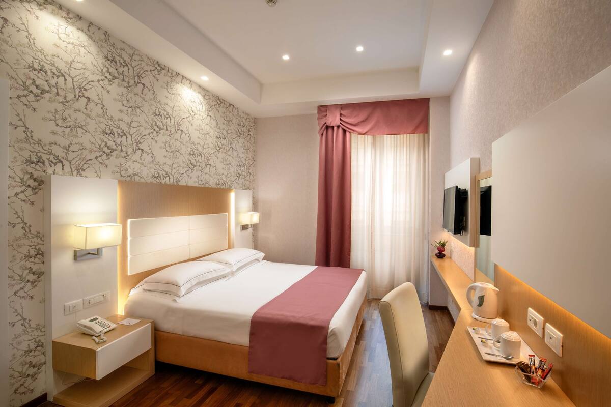 A hotel room with a bed, bedside table, lamp, and a desk with a mirror and a kettle.