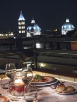 Tavola imbandita per cena con bicchieri di vino, una candela e pane su un balcone con vista su cupole illuminate.