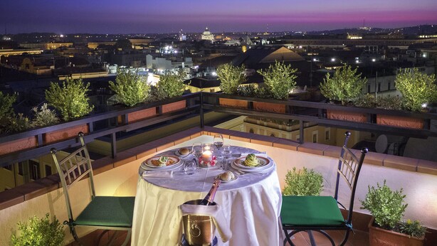 Un área de comedor en la azotea con una mesa preparada para dos, con vistas a un paisaje urbano al atardecer.