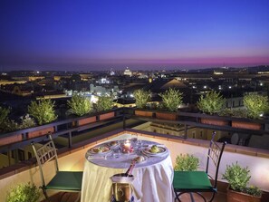 Un área de comedor en la azotea con una mesa preparada para dos, con vistas a un paisaje urbano al atardecer.