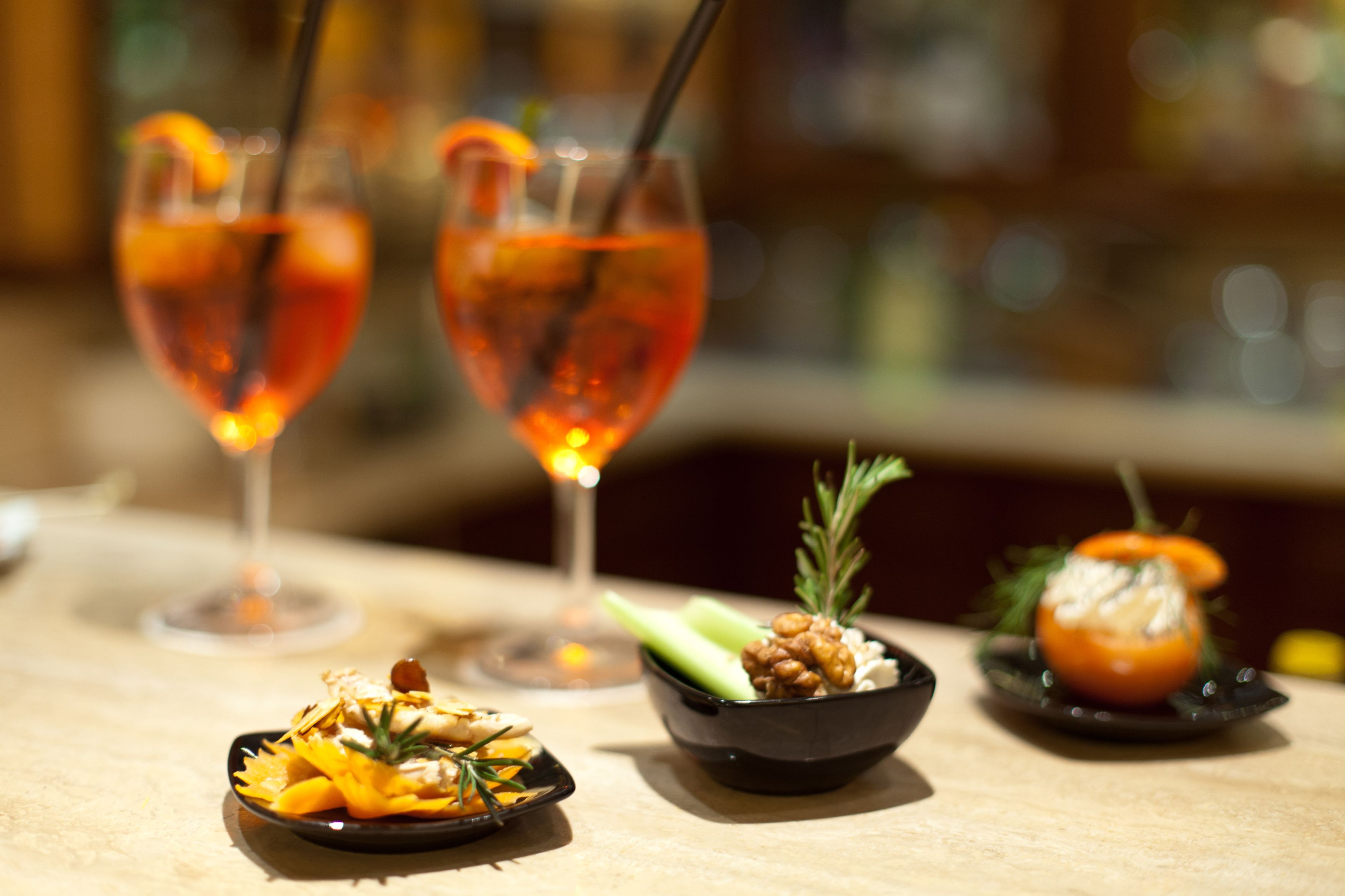 A bar setting with two glasses of a red beverage, a small bowl of nuts and celery, and a plate with a tomato.