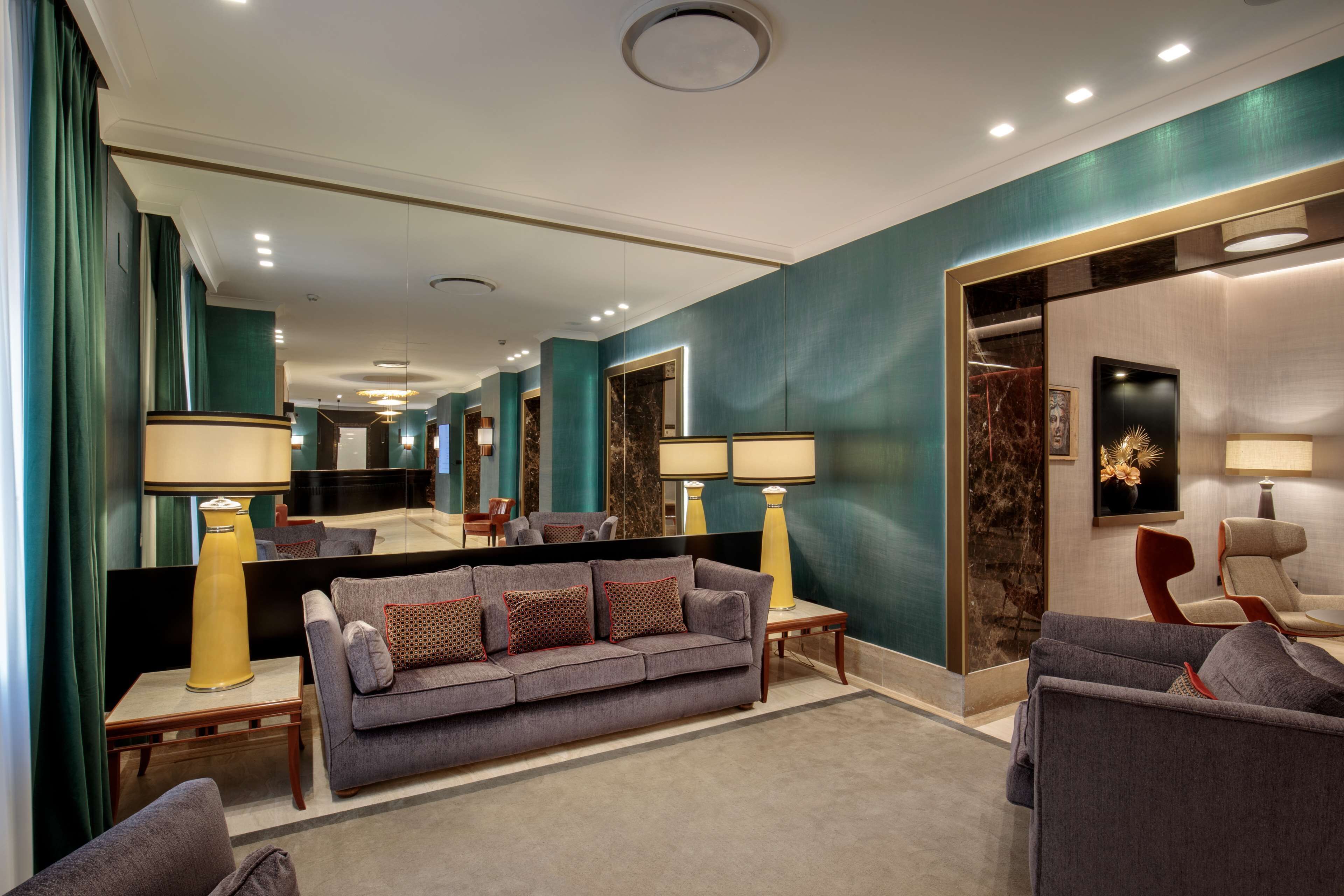 A modern hotel lobby with a grey sofa, a red chair, and a large mirror reflecting the space.