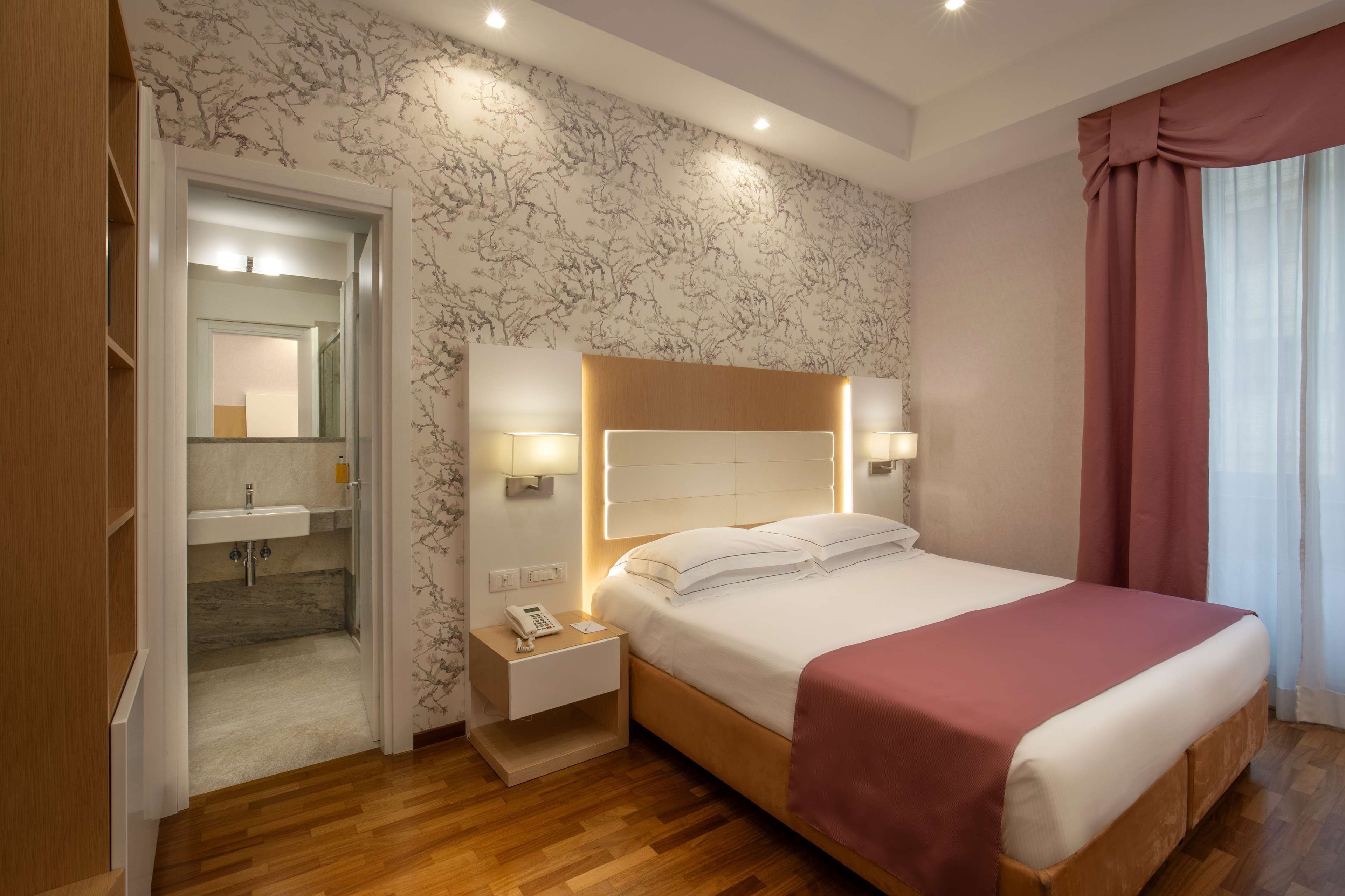 Ein Hotelzimmer mit einem hölzernen Bett, Nachttischlampen, einem Beistelltisch, einem Spiegel und einem durch eine offene Tür einsehbaren Badezimmer.