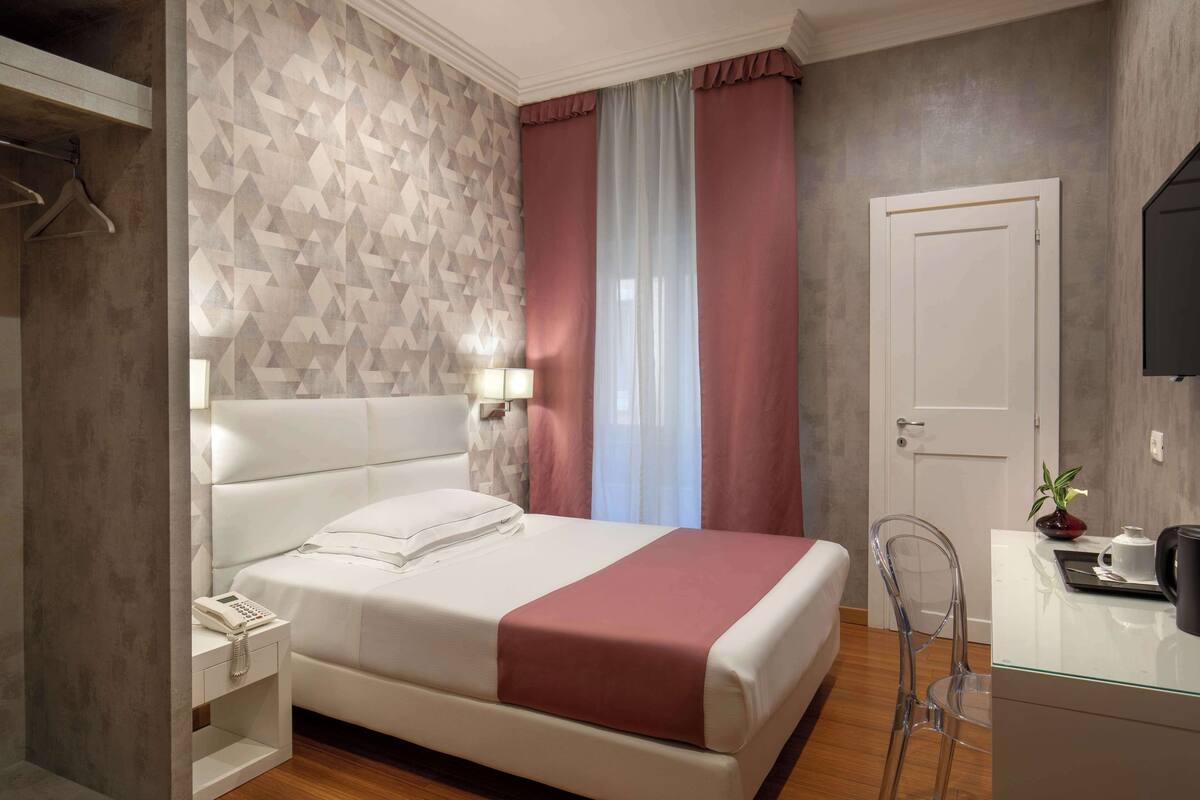 A hotel room with a bed, bedside table, lamp, and a clear chair.