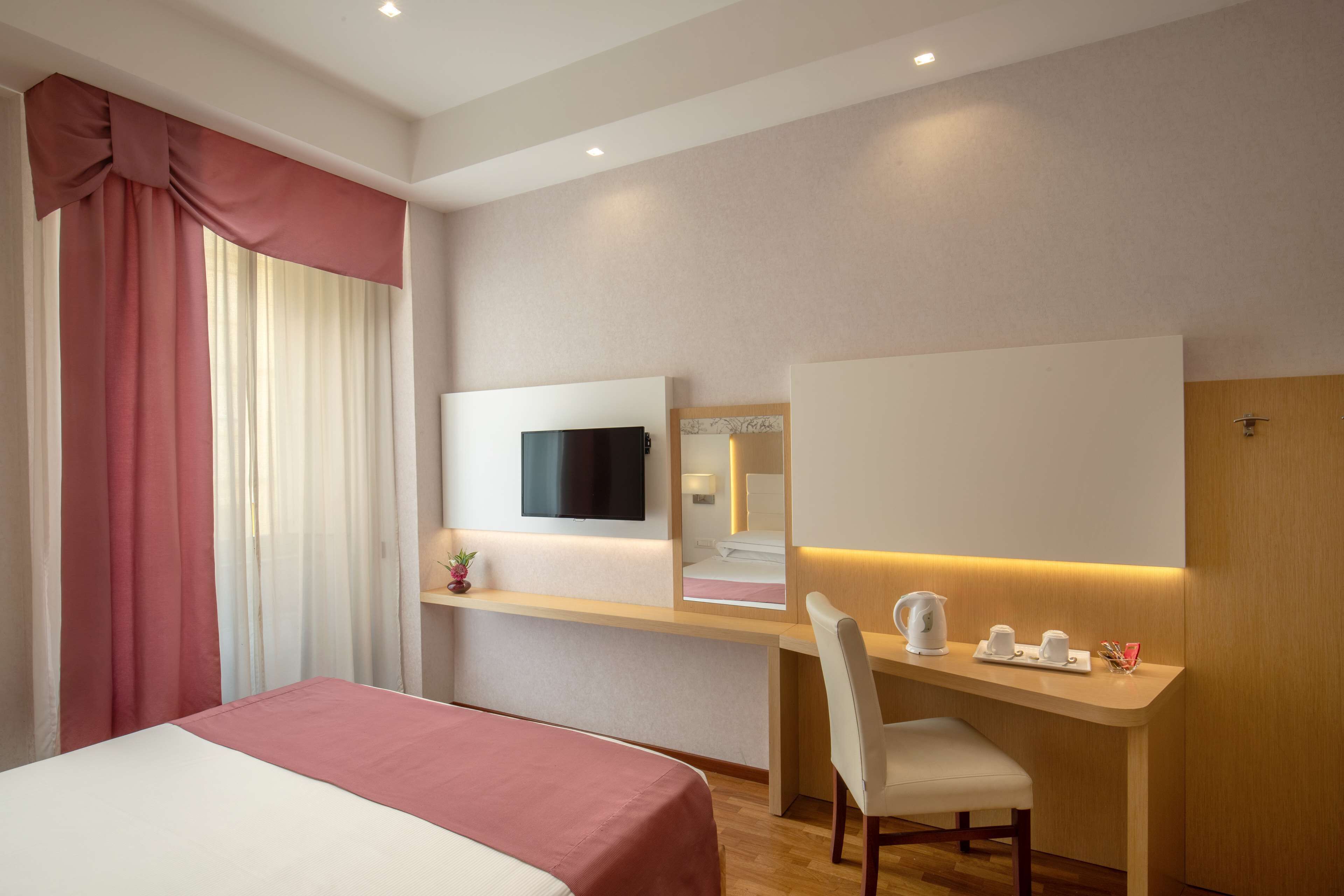 A hotel room with a bed, a desk with a chair, a TV mounted on the wall, and a mirror above the desk.