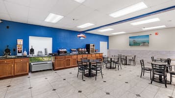 A cafeteria with multiple tables and chairs, a coffee station, and a large window with a view of greenery.