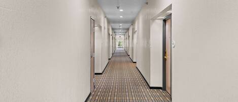 Un pasillo largo y estrecho con puertas a ambos lados, suelo de baldosas y techo sencillo con luces empotradas.