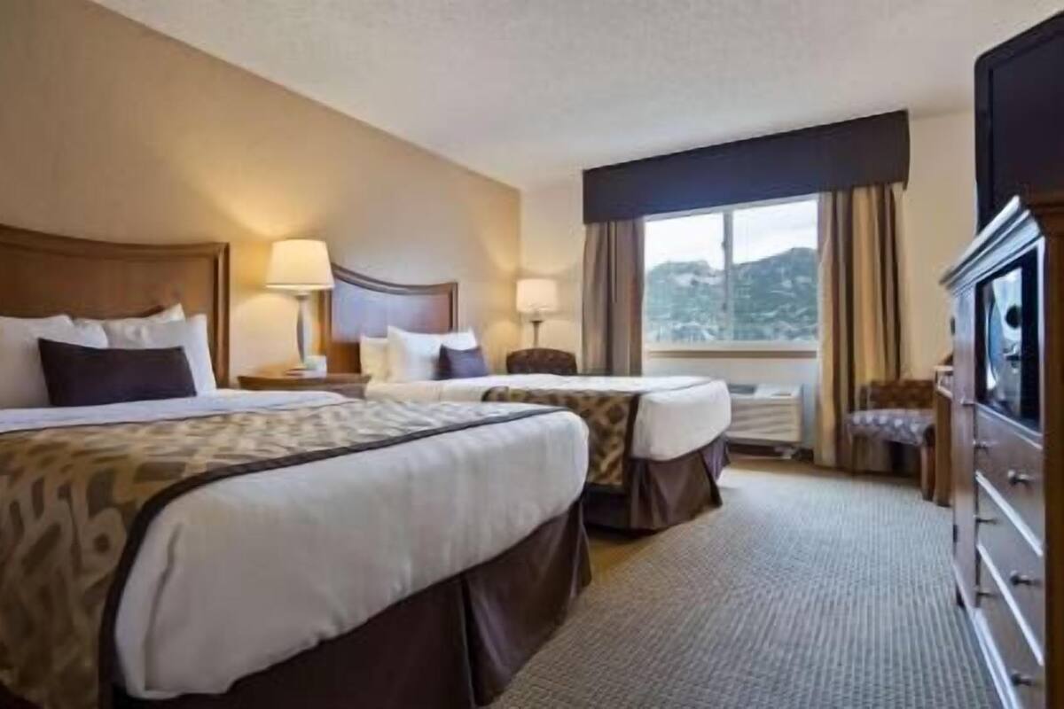 A hotel room with two beds, a dresser, a window with a view, and a lamp on each nightstand.