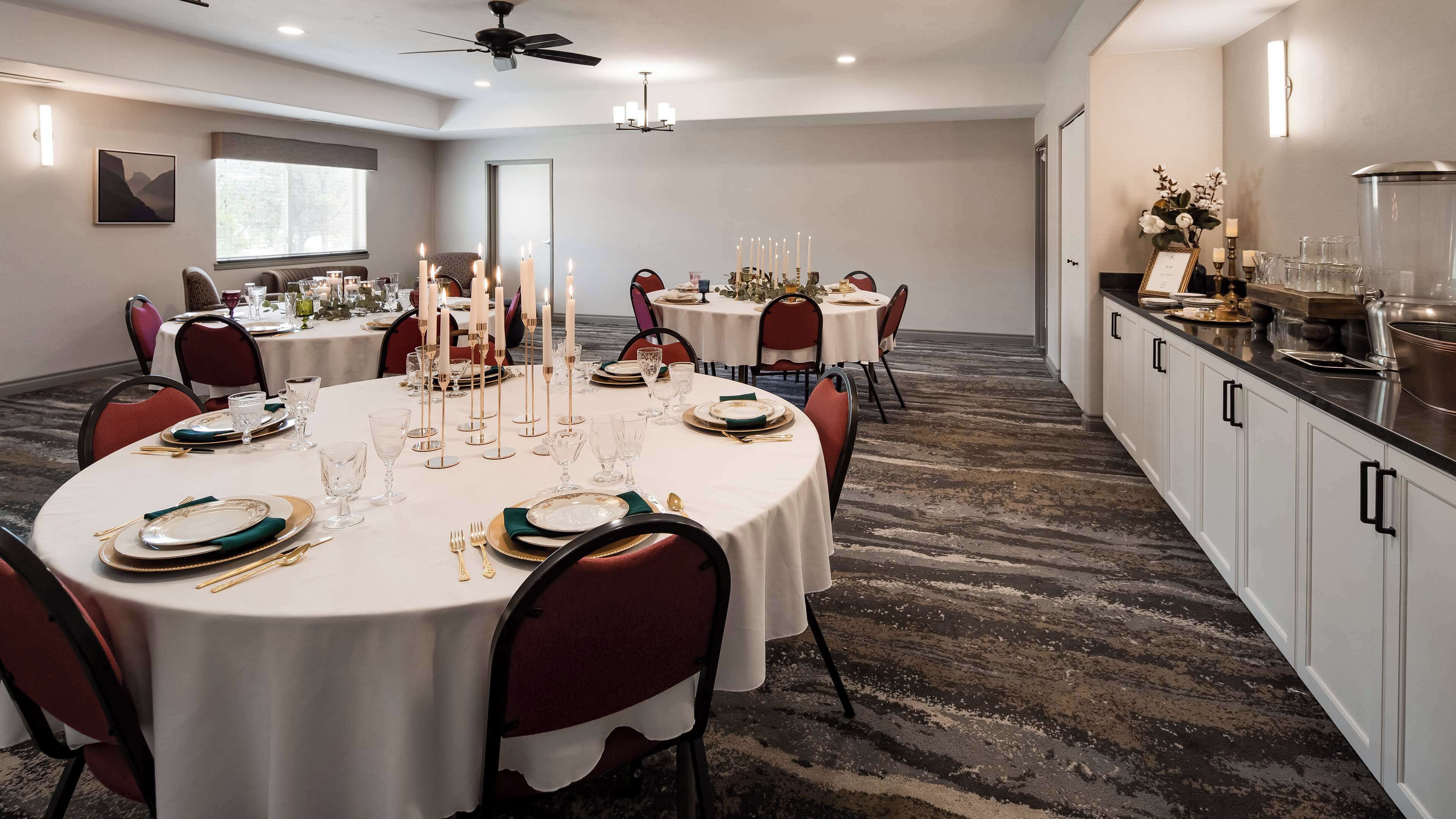 A dining area with round tables set for a meal, featuring white tablecloths, gold and green tableware, and red chairs.