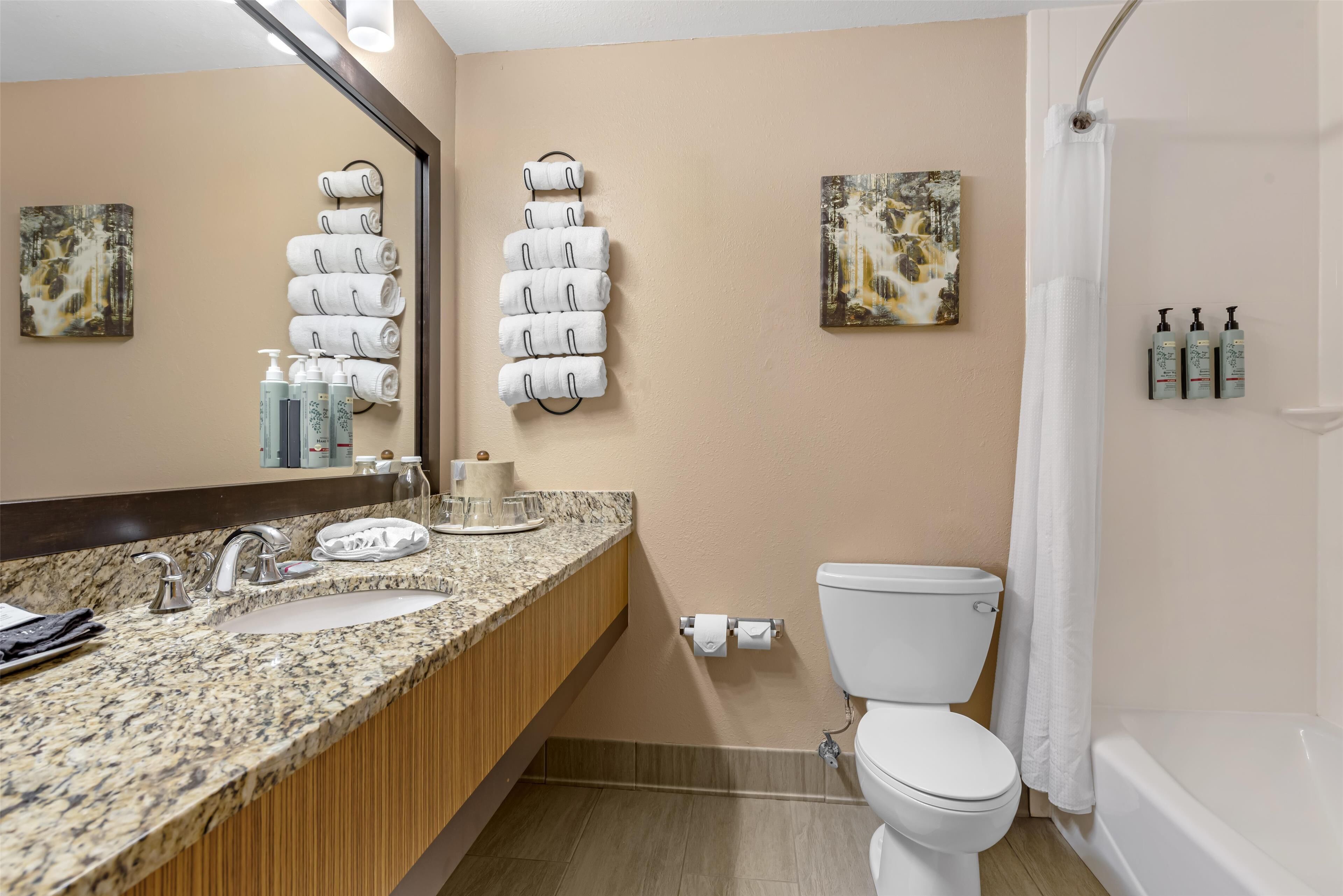 Une salle de bain dotée d’un comptoir en granit, de deux lavabos, de toilettes, d’une douche avec rideau et de serviettes suspendues à des crochets.