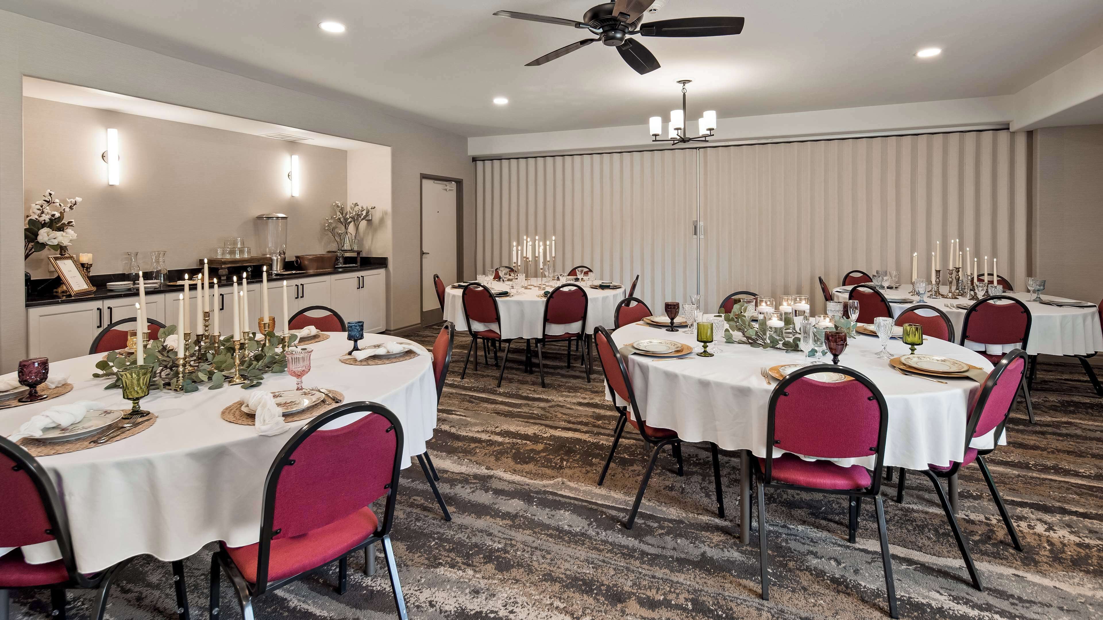 A hotel dining area with round tables set for a meal, featuring white tablecloths, red chairs, and candle centerpieces.