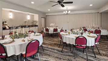 A hotel dining area with round tables set for a meal, featuring white tablecloths, red chairs, and candle centerpieces.