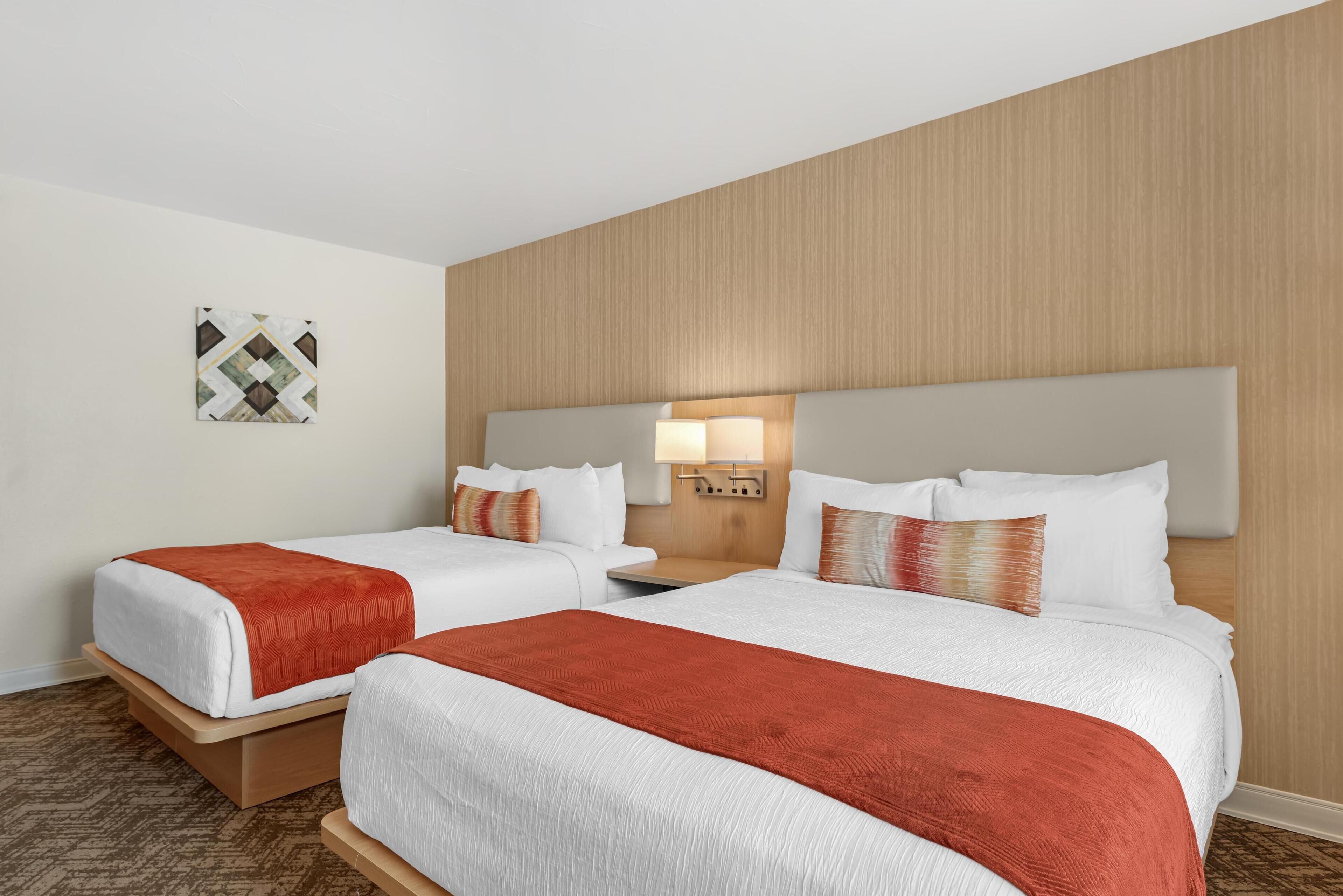 A hotel room with two beds, a nightstand, a lamp, and a wall-mounted artwork.