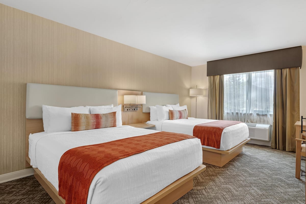 A hotel room with two beds, a large window with curtains, a bedside table with a lamp, and a wall-mounted air conditioner.