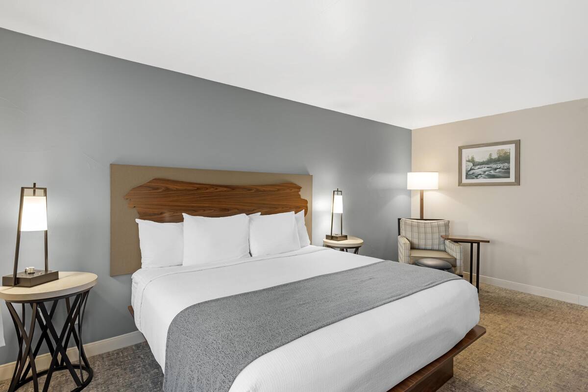 A neatly made bed with a wooden headboard, two bedside lamps, a chair, and a framed picture on the wall.