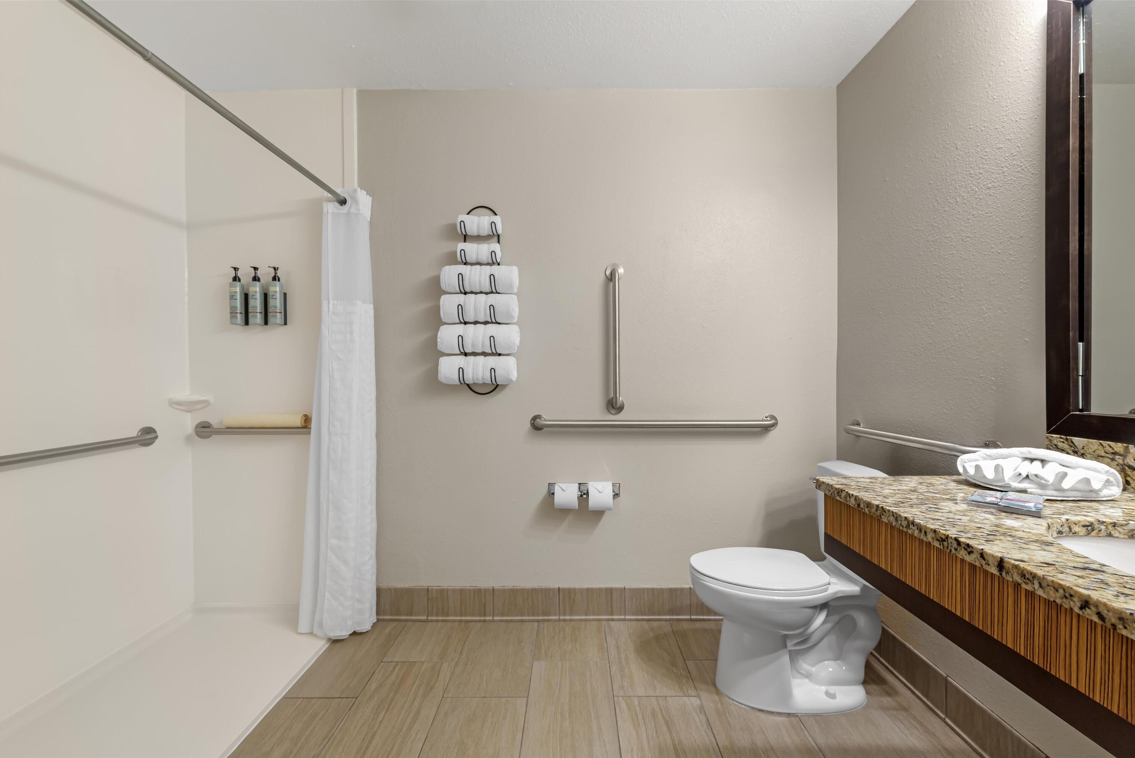 Un baño moderno con ducha, inodoro y lavamanos. Cuenta con barras de apoyo, un perchero para toallas y un dispensador de papel higiénico.