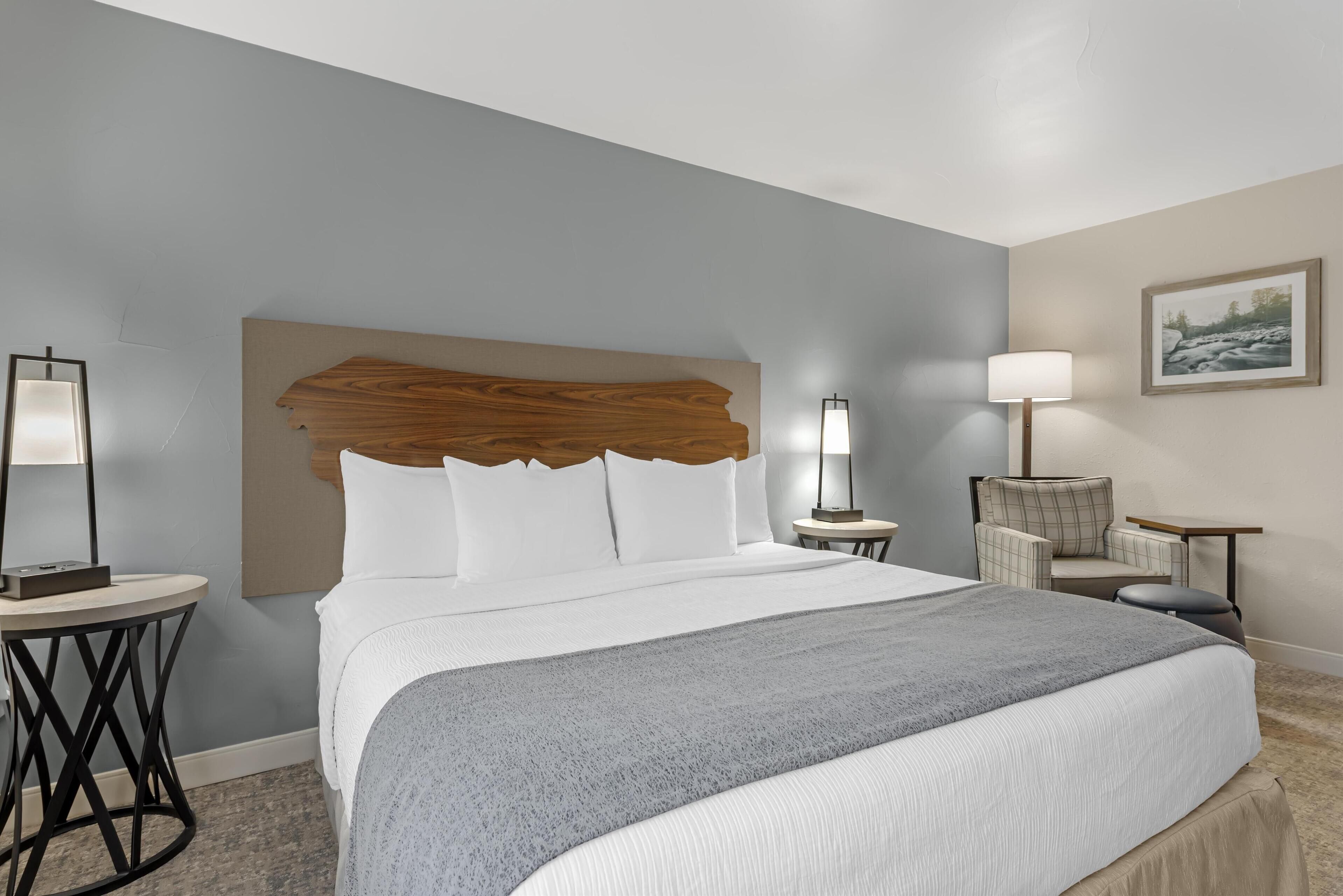 Une chambre d’hôtel avec un grand lit, une tête de lit en bois, une petite table ronde avec une lampe, une chaise et un tableau encadré au mur.