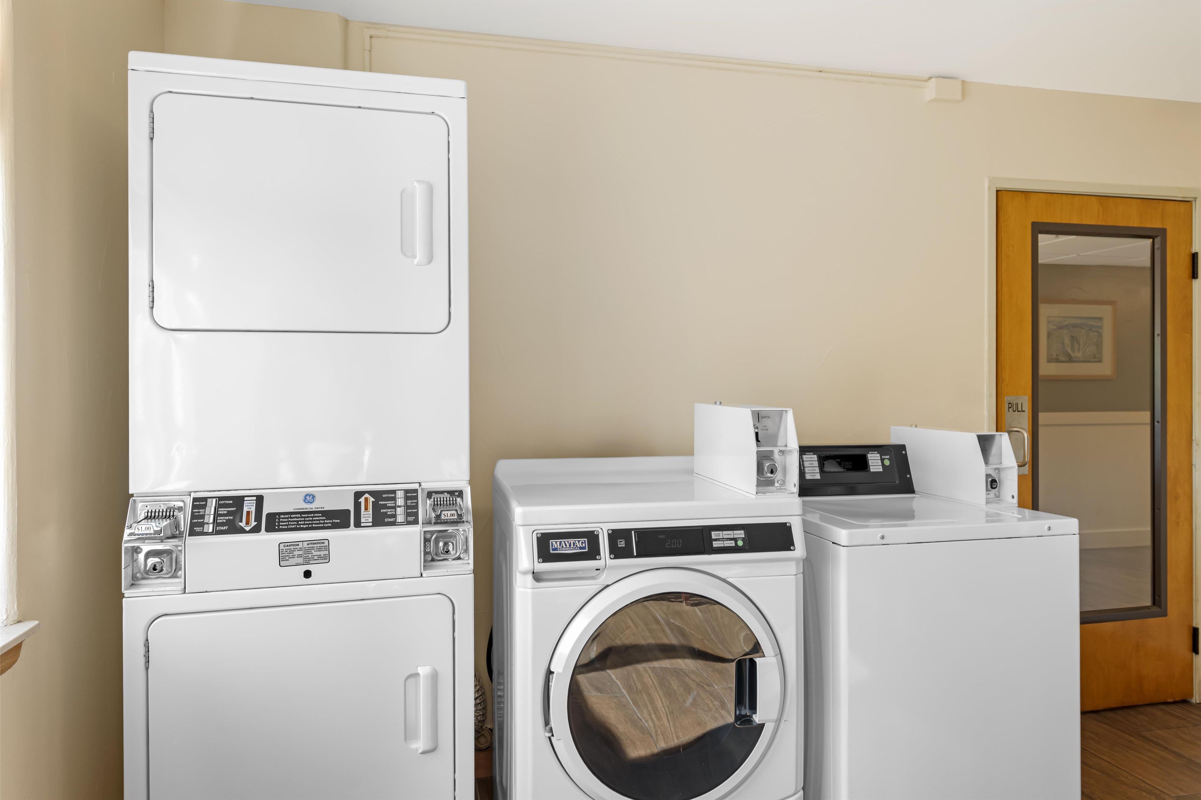 Une buanderie équipée d’une pile de lave-linge et de sèche-linge, une porte avec une indication «  POUSSER » et une prise électrique visible.