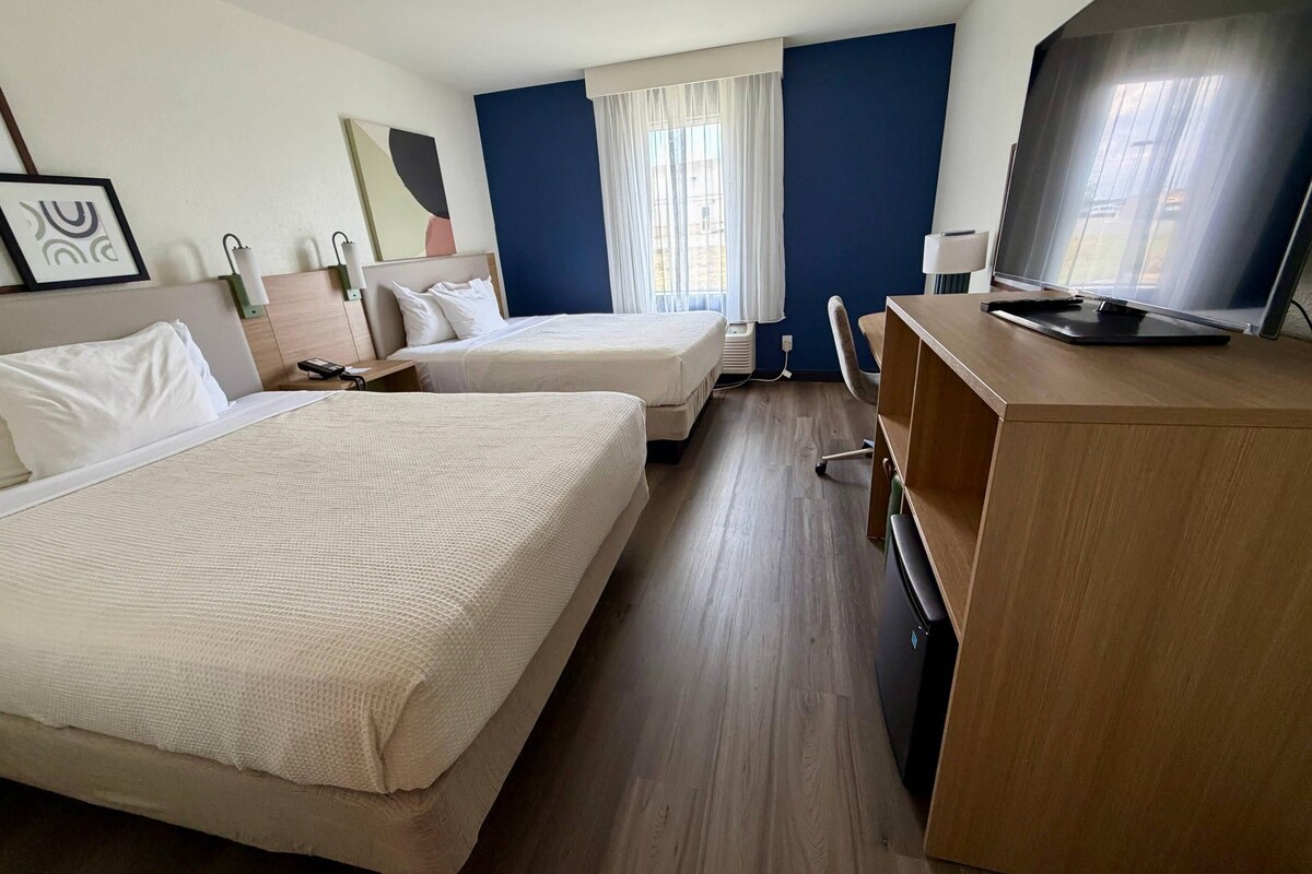 Una habitación de hotel con dos camas, un escritorio, un televisor y una ventana con cortinas.