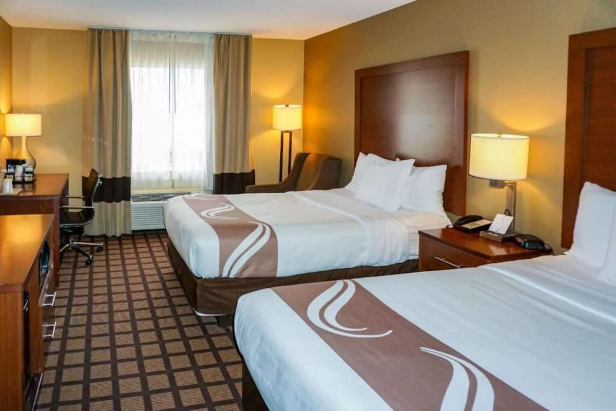 Ein Hotelzimmer mit zwei Betten, einem Schreibtisch, einem Stuhl, einer Lampe und einem Fenster mit Vorhängen.