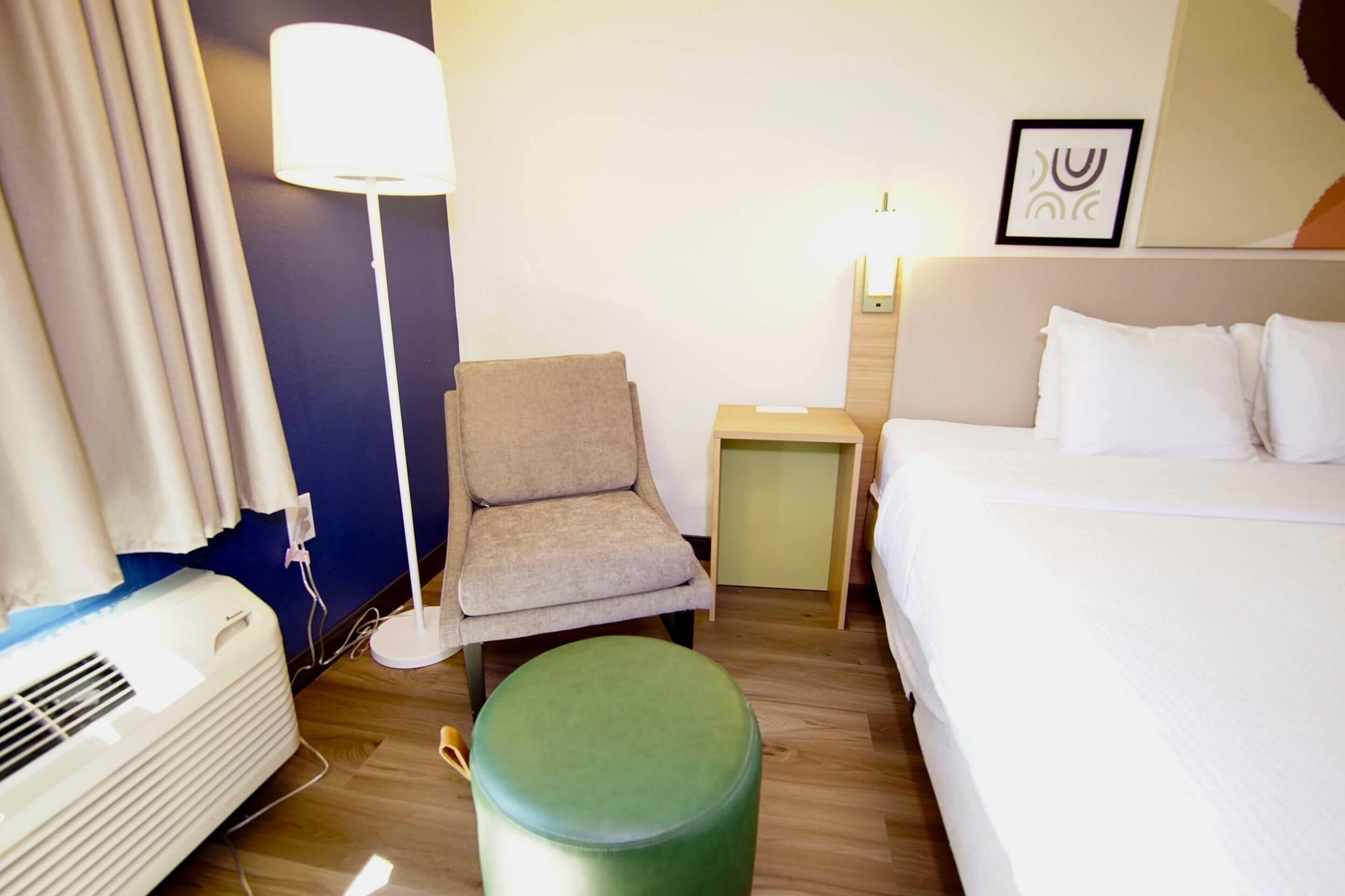 A hotel room with a bed, a chair, a small table, a lamp, and an air conditioning unit.