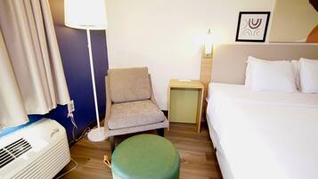 A hotel room with a bed, a chair, a small table, a lamp, and an air conditioning unit.